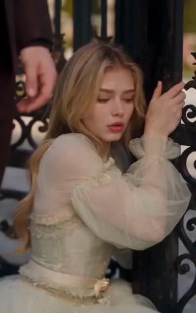 Blonde Erin grips the iron gate of the estate in a pale dress